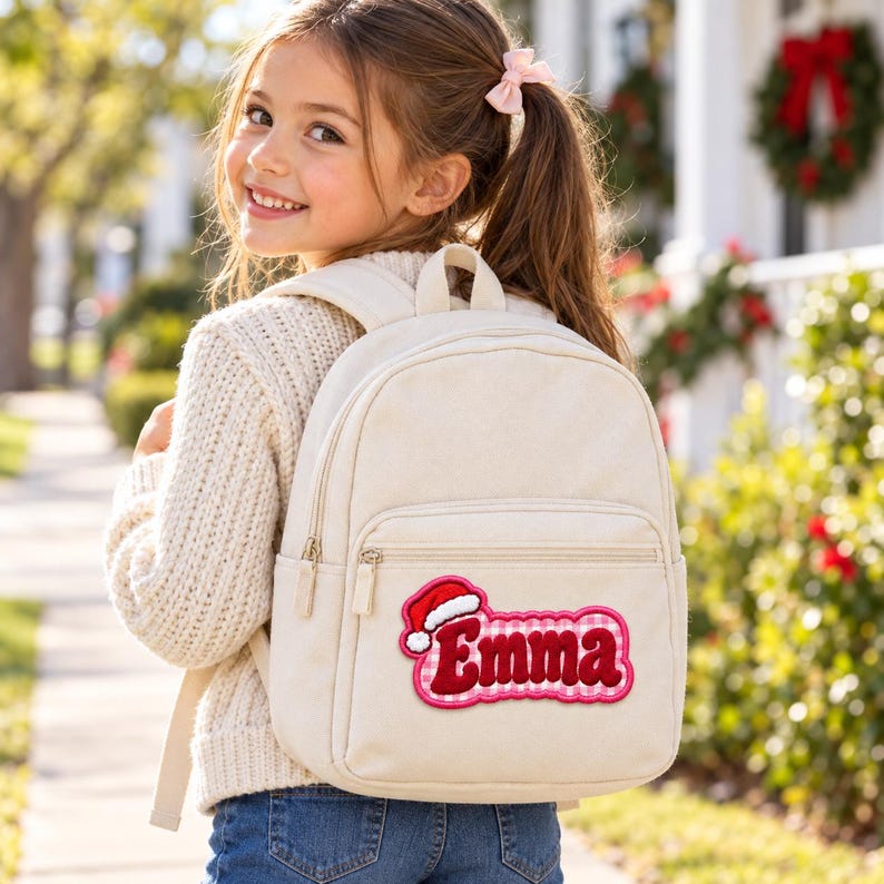 K&ouml;nnte beinhalten: Ein cremefarbener Rucksack aus Segeltuch mit einem personalisierten Aufn&auml;her, der den Namen Emma in roten Buchstaben zeigt, umrandet von einem rosa-wei&szlig; karierten Muster und verziert mit einer kleinen rot-wei&szlig;en Weihnachtsm&uuml;tze.