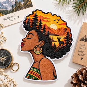 May include: An embroidered patch featuring a profile view of a person with dark skin and red lips, wearing a gold hoop earring. Their hair is styled as a landscape with black pine trees, a golden sun, mountains, a lake, and a black deer silhouette.