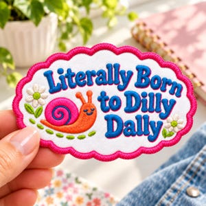May include: An oval embroidered patch with a scalloped hot pink border on a white background. It features a smiling orange snail with a bright pink shell, two white daisies with green leaves, and the blue text Literally Born to Dilly Dally.