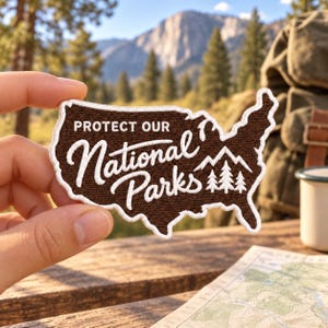 May include: A brown embroidered patch shaped like the map of the United States with a white border. The text reads PROTECT OUR National Parks in white script and block lettering, accompanied by a white graphic of mountain peaks and pine trees.