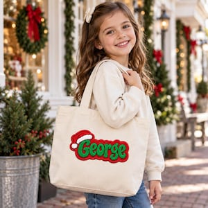 K&ouml;nnte beinhalten: Eine beigefarbene Canvas-Tragetasche mit einem rot-gr&uuml;nen Stickaufn&auml;her, der den Namen George in einer stilisierten Schrift zeigt und mit einer kleinen rot-wei&szlig;en Weihnachtsm&uuml;tze verziert ist. Die Tasche hat zwei Tragegriffe und eine rechteckige Form.