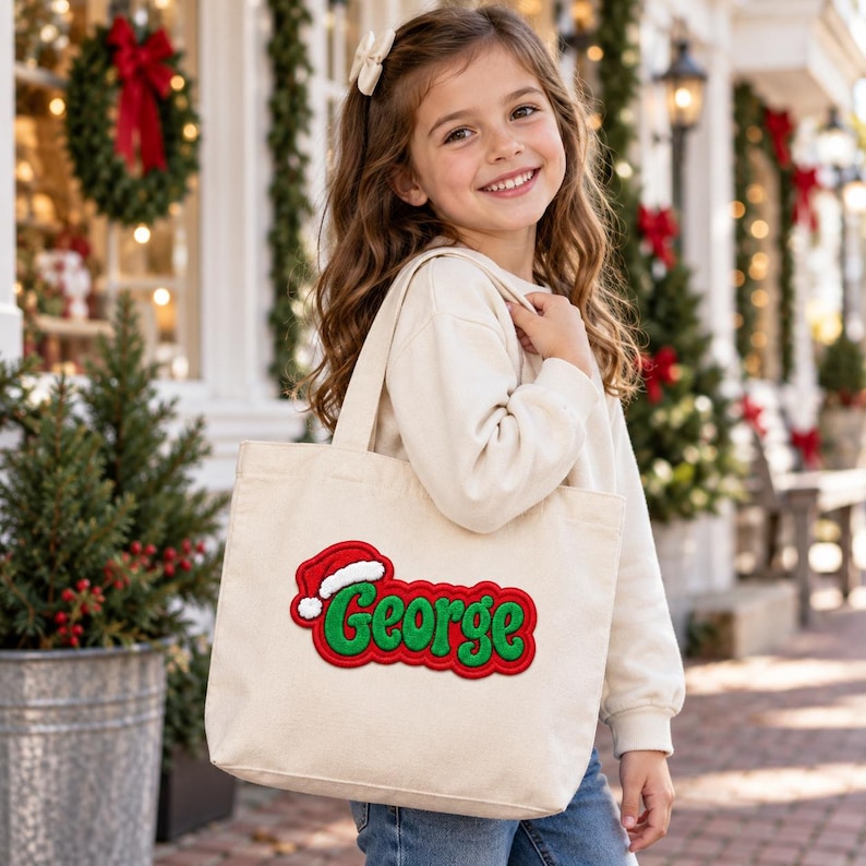 K&ouml;nnte beinhalten: Eine beigefarbene Canvas-Tragetasche mit einem rot-gr&uuml;nen Stickaufn&auml;her, der den Namen George in einer stilisierten Schrift zeigt und mit einer kleinen rot-wei&szlig;en Weihnachtsm&uuml;tze verziert ist. Die Tasche hat zwei Tragegriffe und eine rechteckige Form.