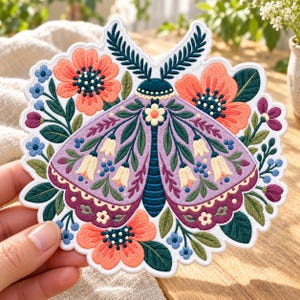 May include: An embroidered patch featuring a central purple moth with cream-colored bell-shaped flowers on its wings, surrounded by vibrant orange and blue flowers, green leaves, and dark teal antennae, all set against a white scalloped border.