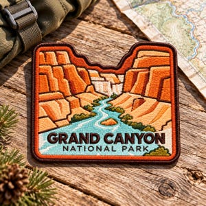 May include: A rectangular embroidered patch featuring a landscape of the Grand Canyon with orange rock formations and a light blue river flowing through the center. The text GRAND CANYON NATIONAL PARK is stitched in dark brown at the bottom.