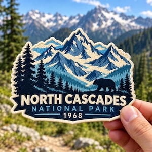 May include: An embroidered patch featuring a mountain landscape with snow-capped peaks, a dark blue forest, and a black bear silhouette. The text reads NORTH CASCADES NATIONAL PARK 1968 in cream and light blue against a dark blue background.
