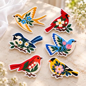 May include: Six embroidered bird patches featuring floral motifs. The collection includes a yellow bird with blue flowers, a red cardinal, a blue bird with white daisies, a blue bird with pink tulips, a red bird with lily of the valley, and a yellow bird with pink flowers.