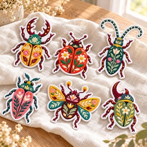 May include: A set of six embroidered patches featuring various beetle and insect shapes. Each patch displays intricate floral and leaf patterns in vibrant colors including red, yellow, green, and pink, with detailed stitching and white borders.