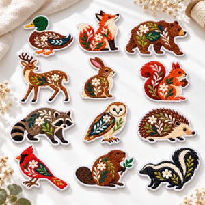 May include: A collection of twelve embroidered iron-on patches featuring woodland animals including a duck, fox, bear, deer, rabbit, squirrel, raccoon, owl, hedgehog, cardinal, beaver, and skunk, each decorated with white flowers and green leaves.