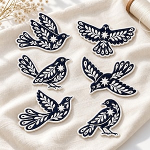 May include: Six dark navy blue embroidered bird patches with white decorative leaf patterns and a central star motif on each wing or body. The patches feature a clean white border and are arranged in two columns on a light beige fabric surface.