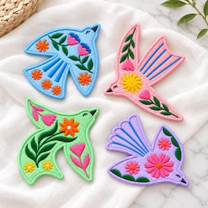 May include: Four embroidered bird-shaped patches in light blue, pink, light green, and lavender. Each patch features colorful floral motifs with orange, pink, and yellow flowers, green leaves, and dark blue or black stitched details on the birds.