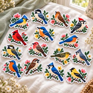 May include: A collection of fifteen embroidered bird patches, each featuring a different bird species perched on a branch with green leaves and colorful flowers, including red, blue, yellow, and brown birds on a white fabric background.
