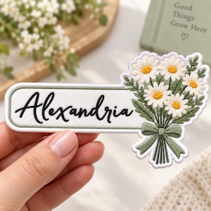 May include: A rectangular embroidered patch with a light green border and white background featuring the name Alexandria in black cursive script. Attached to the right side is an embroidered bouquet of white daisies with yellow centers and green stems tied with a green bow.