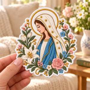 May include: An embroidered patch featuring a profile of the Virgin Mary wearing a white veil with gold star accents and a blue robe, surrounded by a decorative border of white lilies, pink roses, and small blue flowers on a white background.