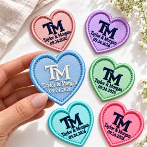 May include: Six heart-shaped embroidered patches in various pastel colors including pink, purple, light blue, green, and teal. Each patch features the monogram TM, the names Taylor & Morgan, and the date 09.24.2026 stitched in dark blue or white thread.