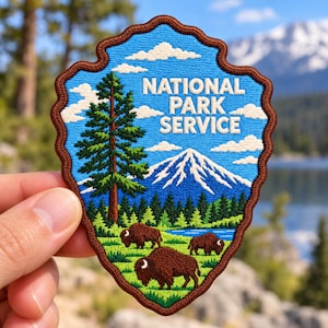 May include: An embroidered patch with a brown scalloped border featuring a blue sky, white clouds, a tall green pine tree, a snow-capped mountain, a blue lake, and three brown bison grazing on green grass with the text NATIONAL PARK SERVICE.