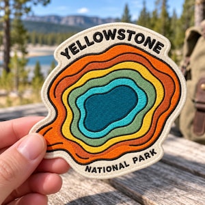 May include: An embroidered patch featuring a topographic map design with concentric rings in shades of orange, yellow, green, and blue. The text YELLOWSTONE is arched at the top and NATIONAL PARK is at the bottom in black lettering.