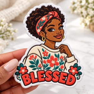 May include: An embroidered patch featuring a person with dark skin and curly hair wearing a red floral headband and gold hoop earrings. The person wears a cream-colored sweater with red and green floral patterns. The word BLESSED is in red letters.