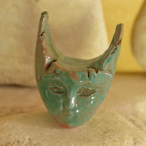 Peut inclure: Un petit ornement de visage en céramique sculpté à la main avec une glaçure brillante vert mousse. L'objet présente deux protubérances pointues en forme de cornes au sommet, des yeux en amande, un nez et une bouche subtils, avec de l'argile brun-rouge.