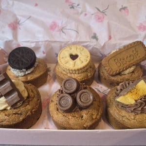 May include: A selection of six cupcakes in a pink box, each decorated with various toppings. These include an Oreo, a custard cream biscuit, and a Biscoff biscuit. Other cupcakes are topped with chocolate.