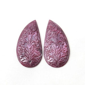 Puede incluir: Dos piedras preciosas de rubí en forma de lágrima con intrincados patrones florales tallados a mano. Cada piedra de color rojo intenso muestra un diseño simétrico de tallos, hojas y flores grabado en la superficie, con un acabado pulido.