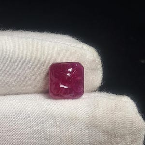 May include: A square-shaped, deep red gemstone featuring a carved floral pattern on its surface. The stone has a smooth, polished finish with a central circular detail surrounded by petal-like engravings, displaying a rich, translucent crimson hue.