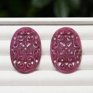 Puede incluir: Dos cabujones de piedra preciosa de forma ovalada en color magenta intenso, con intrincados tallados de filigrana calada que muestran un motivo floral central rodeado de patrones simétricos en espiral. Piedras pulidas y translúcidas.