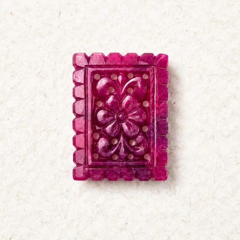 Op de afbeelding: Een rechthoekige, diep magentakleurige edelsteen hanger met een ingewikkeld, uitgesneden bloemmotief in het midden. Het stuk is omlijst door een geschulpte, ingekeepte rand en bevat kleine, ronde uitsparingen rondom het centrale bloemendesign.