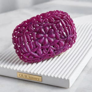 Puede incluir: Una talla de piedra preciosa de color magenta intenso con forma ovalada que presenta un intrincado patrón floral y de hojas caladas. La pieza descansa sobre un soporte de exhibición blanco, rectangular y acanalado con una placa de metal dorado grabada con el texto OUR luxury.