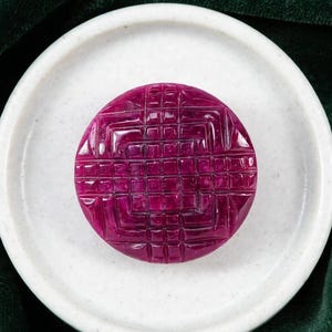 May include: A circular, deep magenta gemstone button featuring a carved, grid-like geometric pattern across its surface. The button has a polished, reflective finish and is centred on a smooth, off-white, circular ceramic dish.