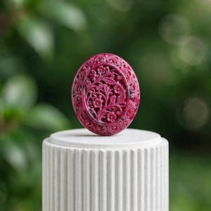以下が含まれることがあります： 深いマゼンタ色の楕円形のペンダントで、咲き誇る花々と絡み合う蔓の複雑な彫刻レリーフが施されています。滑らかで光沢のある外縁と、質感のある詳細な花の中心部が特徴の装飾品です。