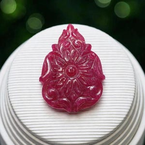 Puede incluir: Un colgante de piedra preciosa de color rojo intenso y translúcido, tallado con un motivo floral simétrico en forma de lágrima. La superficie presenta intrincados patrones de pétalos en relieve que irradian desde un punto central circular.