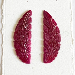 May include: Two deep magenta, crescent-shaped carved gemstone pieces featuring a detailed floral and leaf motif. Each piece displays a central flower surrounded by textured foliage, with a polished, glossy finish and symmetrical, curved edges.