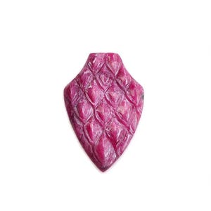 May include: A shield-shaped pendant carved from a deep magenta-pink gemstone. The surface features a repeating, raised diamond-pattern texture that resembles scales, with a flat top edge and a pointed bottom tip, showcasing natural stone variations.