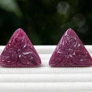 Pode incluir: Duas pedras preciosas triangulares de cor magenta profundo com um padrão floral esculpido nas superfícies frontais. As pedras têm um acabamento liso e polido e estão posicionadas lado a lado, exibindo detalhes de pétalas simétricos.