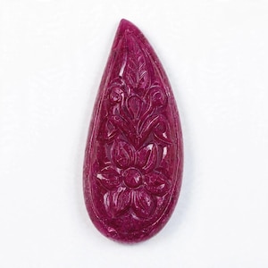 May include: A teardrop-shaped gemstone in a deep magenta colour, featuring an intricate hand-carved floral design on its surface. The carving displays a central flower with petals and leaves extending upwards, creating a textured relief pattern.