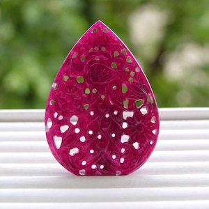 May include: A teardrop-shaped pendant carved from deep magenta stone featuring an intricate openwork design of swirling floral patterns and leaves. The surface displays a polished finish with visible natural veining throughout the gemstone.