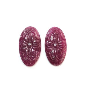 Op de afbeelding: Twee ovaalvormige, diep magenta gekleurde edelstenen met een uitgesneden bloemmotief op hun oppervlak. Elke steen heeft een gladde, gepolijste afwerking met symmetrische bloemblaadjes die vanuit het midden uitstralen, wat een gedetailleerd reliëf creëert.