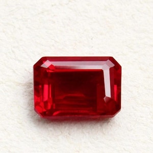 May include: A vibrant, deep red gemstone cut into a rectangular emerald shape. The stone features precise, multi-faceted edges that reflect light, creating a clear, polished surface with varying shades of crimson and scarlet throughout the gem.