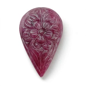 May include: A teardrop-shaped gemstone in a deep magenta-red hue, featuring an intricate, hand-carved floral design on its surface. The carving displays a central flower with symmetrical petals and decorative scrollwork extending toward the pointed base.