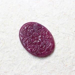 Könnte beinhalten: Ein ovaler, tief magentafarbener Edelstein-Cabochon mit einem detaillierten, geschnitzten Blumenmuster auf der Oberfläche. Der Stein hat eine glatte, polierte Oberfläche mit erhabenen Relieflinien, die die Blütenblätter und Blätter des Designs definieren.