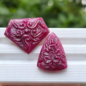 以下が含まれることがあります： 複雑な花のレリーフ模様が彫られた、深いマゼンタ色の宝石カボション2点。1点は五角形で、もう1点は角が丸い三角形です。どちらの石も磨き上げられた光沢のある仕上げで、細部まで精巧な彫刻が施されています。