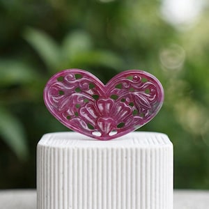 Peut inclure: Un pendentif en forme de cœur sculpté dans une pierre magenta profonde, présentant des motifs floraux et végétaux ajourés complexes. La pièce est posée sur un support cylindrique blanc à nervures verticales, révélant sa texture lisse.