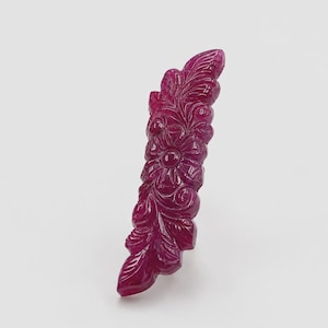Pode incluir: Uma gema alongada de cor magenta profunda com intrincados motivos florais e de folhas esculpidos. A superfície da pedra é polida e apresenta um trabalho em relevo detalhado, exibindo uma flor central rodeada por padrões de folhagem.