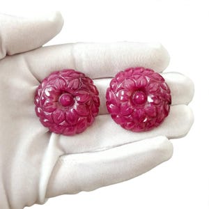 Könnte beinhalten: Zwei kreisförmige, tiefrosa Edelstein-Cabochons mit einem detaillierten, geschnitzten Blumenmuster, bei dem geschichtete Blütenblätter von einem zentralen, glatten, erhabenen Punkt ausgehen. Die Steine haben eine polierte, glänzende Oberfläche.