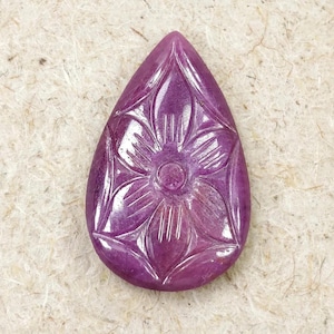 May include: A teardrop-shaped gemstone in a deep magenta-purple hue, featuring a hand-carved floral design with a central circular element and six radiating petals. The surface of the stone is polished and smooth, highlighting the natural texture.