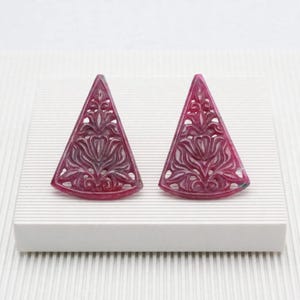 Puede incluir: Un par de pendientes triangulares hechos de piedra color magenta intenso, con intrincados tallados calados de motivos florales y de hojas. Los pendientes tienen una base plana y una parte superior puntiaguda, mostrando un diseño simétrico.