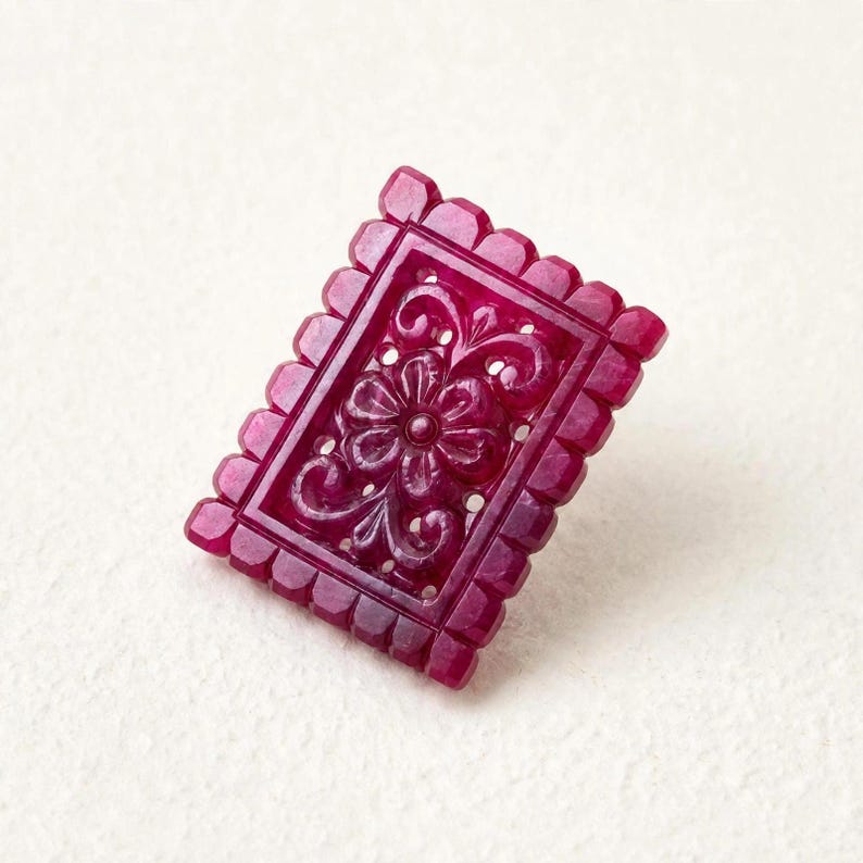 Op de afbeelding: Een rechthoekig, diep magentakleurig edelsteensnijwerk met een ingewikkeld, verhoogd bloemmotief in het midden. Het stuk is omlijst door een geschulpte, ingekeepte rand die langs alle vier de zijden loopt en een getextureerde, decoratieve rand vormt.