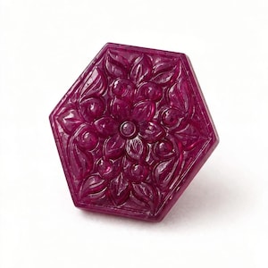 Puede incluir: Un botón de piedra preciosa de forma hexagonal en color magenta intenso, con un patrón floral tallado y simétrico de pétalos y hojas en relieve. La superficie es pulida y translúcida, mostrando detalles intrincados en su forma.
