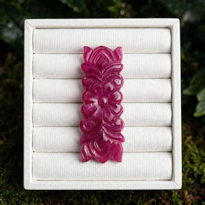 May include: A rectangular, deep magenta-colored gemstone carving featuring a symmetrical, intricate floral and leaf relief pattern. The piece is centered on a white, multi-slotted jewelry display insert, showcasing a polished, glossy surface texture.