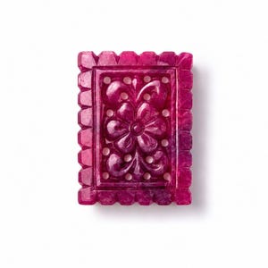 May include: A rectangular, deep magenta-colored gemstone carving featuring an intricate, openwork floral motif at its center. The piece is framed by a scalloped, notched border, showcasing a polished, translucent surface with natural mineral inclusions.